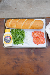 A small tray containing French bread, a container of mayo, sliced onions, and sliced tomatoes for assembling fried oyster poboys.
