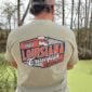 Outdoors, a person wears a “Louisiana Crawfish Company Established 1985” shirt, perfect for Boiled IQF Crawfish Party Packs.