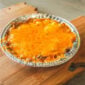 Rachael’s 8oz Shrimp Au Gratin in a foil pan, topped with melted, browned cheese on a wooden cutting board.
