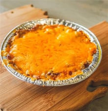 Rachael's Shrimp Au Gratin 8oz Rachael’s 8oz Shrimp Au Gratin in a foil pan, topped with melted, browned cheese on a wooden cutting board.