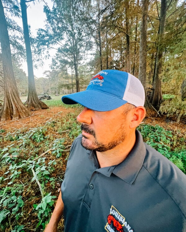 A man wearing an LA Crawfish Co. mesh hat stands in a wooded area.