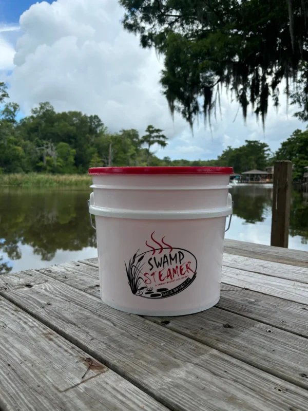 A white plastic bucket with a Swamp Bucket Steamer Attachment on a wooden dock overlooking a bayou.