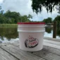 A white plastic bucket with a Swamp Bucket Steamer Attachment on a wooden dock overlooking a bayou.