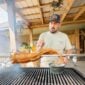 A man grills a bone-in whole skinned alligator tail on a rotisserie over a charcoal grill.