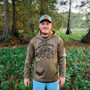A man outdoors wearing a baseball cap and a light brown LA Crawfish Co. Sport-Tek hoodie with a crawfish graphic.