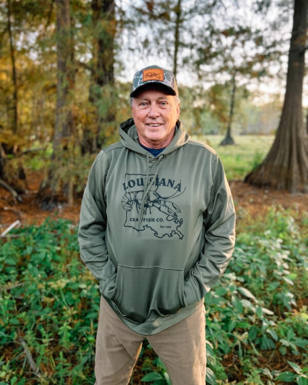 green hoodie A man in a camo cap and olive green Sport-Tek hoodie with a Louisiana Crawfish Co. graphic stands outdoors.