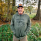 A man in a camo cap and olive green Sport-Tek hoodie with a Louisiana Crawfish Co. graphic stands outdoors.