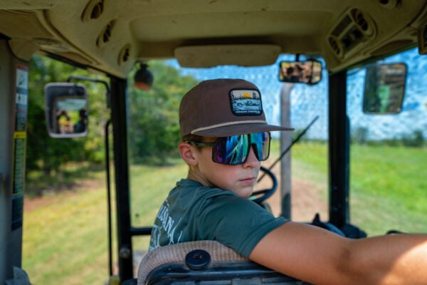 A boy in sunglasses and a brown LA Crawfish Co. Boat Patch Grandpa Hat, sitting inside a tractor cab.
