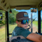 A boy in sunglasses and a brown LA Crawfish Co. Boat Patch Grandpa Hat, sitting inside a tractor cab.