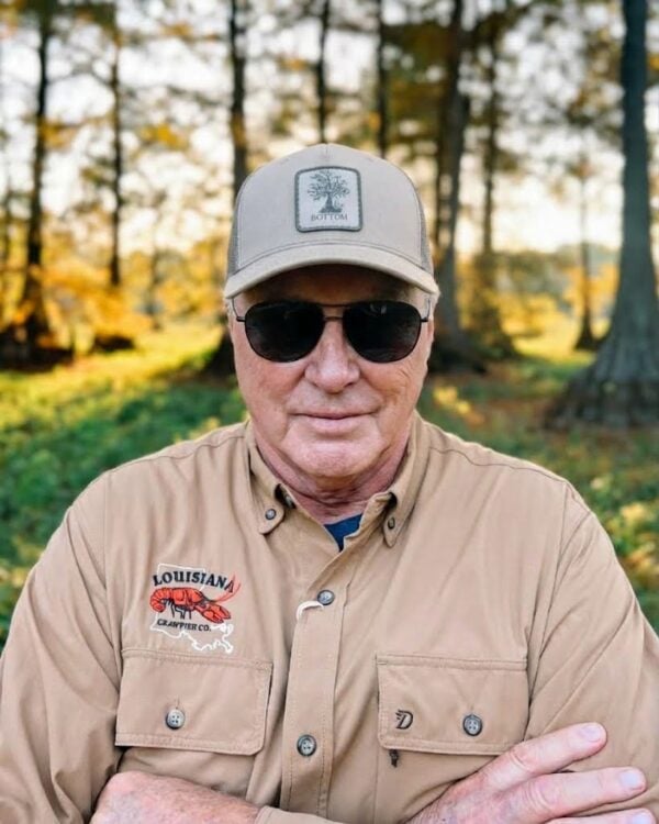 A man wearing sunglasses and a tan Swamp Bottom patch hat with a tree graphic, standing in the woods.