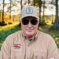 A man wearing sunglasses and a tan Swamp Bottom patch hat with a tree graphic, standing in the woods.