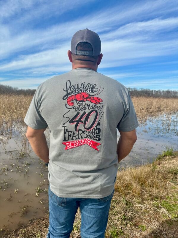 A man in a cap stands by a wet field, wearing a gray 2025 LA Crawfish Co. 40th Anniversary shirt celebrating Louisiana seafood.