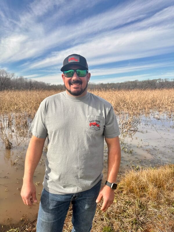 IMG_3627__17402.1737057400.1280.1280.jpg A man in sunglasses and a cap stands by a flooded field, wearing jeans and a 2025 LA Crawfish Co. anniversary shirt.