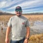 A man in sunglasses and a cap stands by a flooded field, wearing jeans and a 2025 LA Crawfish Co. anniversary shirt.