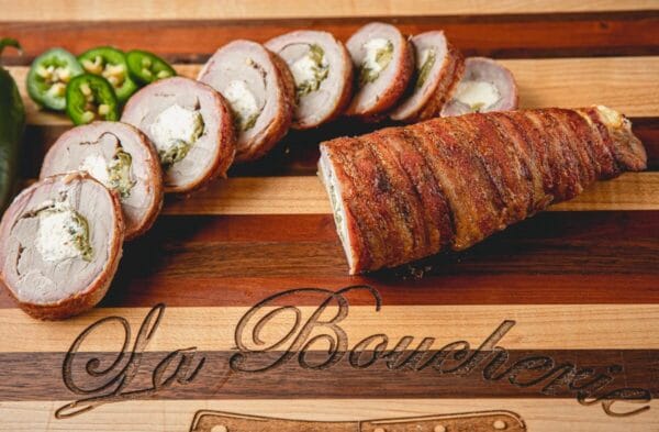 Sliced, bacon-wrapped pork tenderloin stuffed with cream cheese and jalapeño on a board, with extra jalapeño slices nearby.