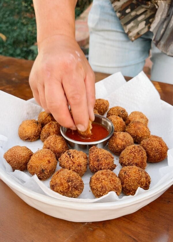 boulettes prepared A hand dips a Shrimp and Crab Boulette into red sauce, served alongside a platter of fried appetizers.