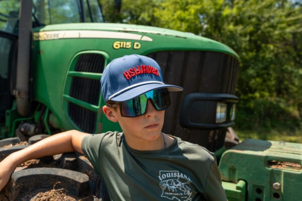 A person wearing reflective sunglasses and a blue backwards Crawfish hat stands in front of a green tractor.