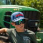 A person wearing reflective sunglasses and a blue backwards Crawfish hat stands in front of a green tractor.