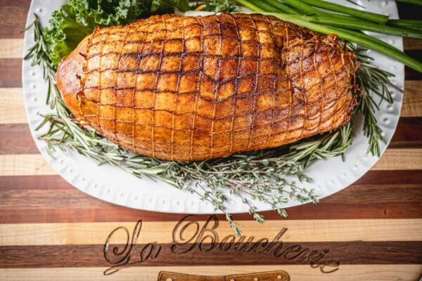 A cooked 5 lb La Boucherie Turducken Roll sits on a white platter garnished with herbs atop a wooden table.