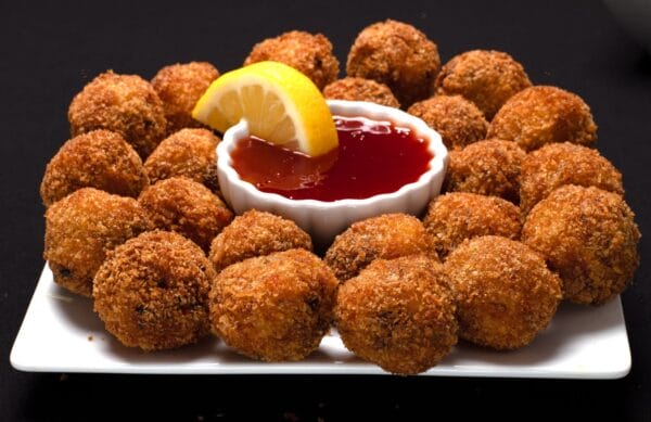 boulette2__77570.1754322102.1280.1280.jpg Shrimp and crab boulettes on a square plate, surrounding a bowl of red sauce and a lemon wedge.