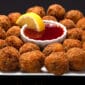 Shrimp and crab boulettes on a square plate, surrounding a bowl of red sauce and a lemon wedge.