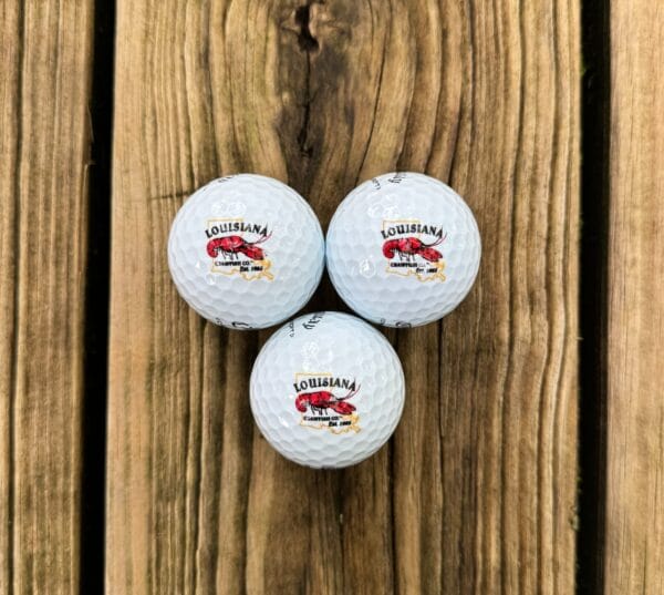 Three white LA Crawfish Co. golf balls with Louisiana logos rest on a wooden surface, ideal for Louisiana fans.