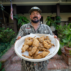 A man stands outside near a house, holding a plate of frog legs, with an American flag waving nearby.