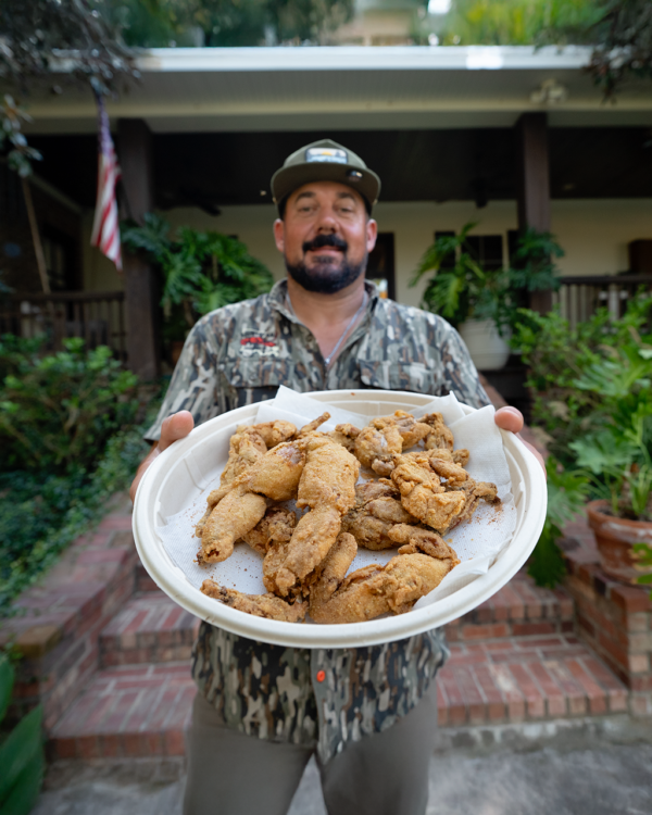 frog_legs_fried__95369.1756850317.1280.1280.png A man in camouflage holds a plate of 2/4 ct, 5 lb frog legs outside, near brick steps, a house, and greenery.