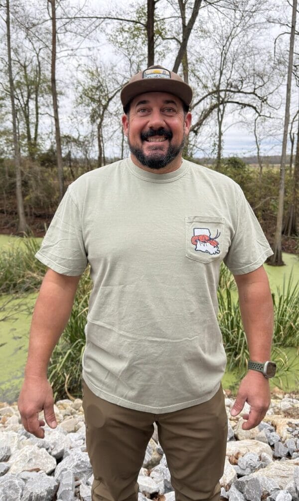 2026_Front__37383.1765908706.1280.1280.JPG A man in a 2026 LA Crawfish Co T-shirt smiles by a pond, enjoying Louisiana crawfish and Cajun food under shady trees.