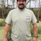 A man in a 2026 LA Crawfish Co T-shirt smiles by a pond, enjoying Louisiana crawfish and Cajun food under shady trees.