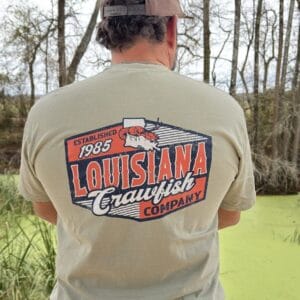 Near a pond, a person wearing a 2026 LA Crawfish Co T-shirt enjoys authentic Louisiana crawfish tradition.
