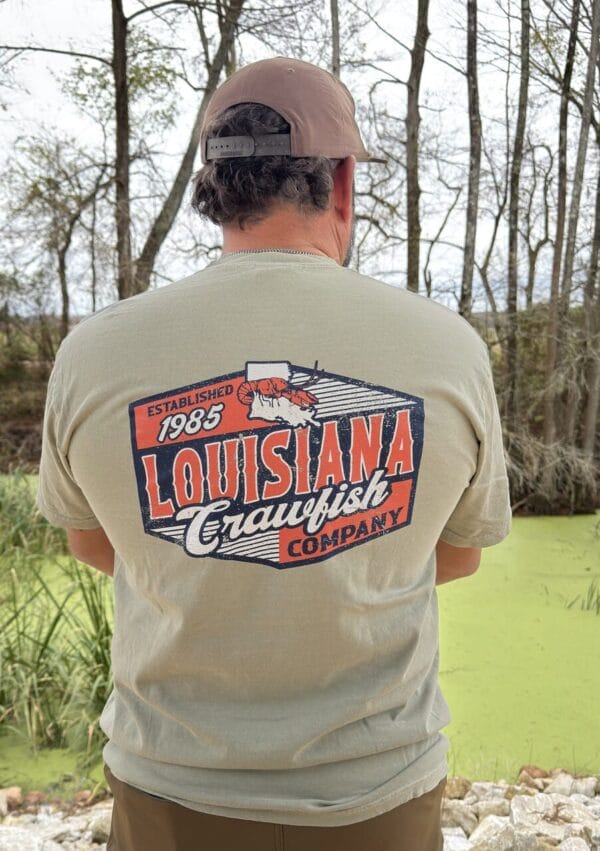 Near a pond, a person wearing a 2026 LA Crawfish Co T-shirt enjoys authentic Louisiana crawfish tradition.
