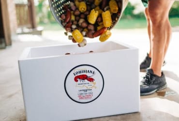 Someone pours corn and boiled food from a pot into a white cooler labeled “Louisiana Seafood” for Cajun cuisine.