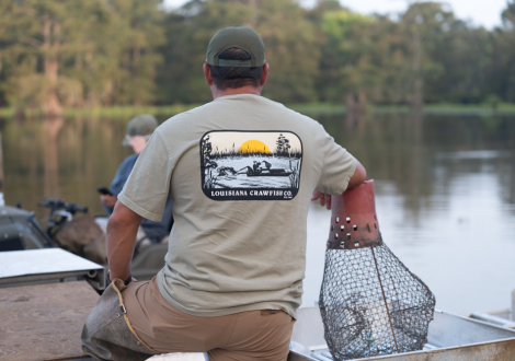 A man in a “Louisiana Crawfish Co.” shirt sits by a lake with a metal basket, capturing true Louisiana crawfish tradition.