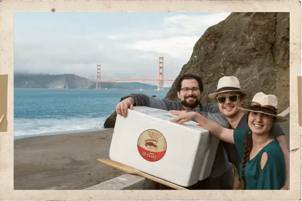 Three people with a cooler stand on a beach, Golden Gate Bridge behind them, set to enjoy Louisiana crawfish.