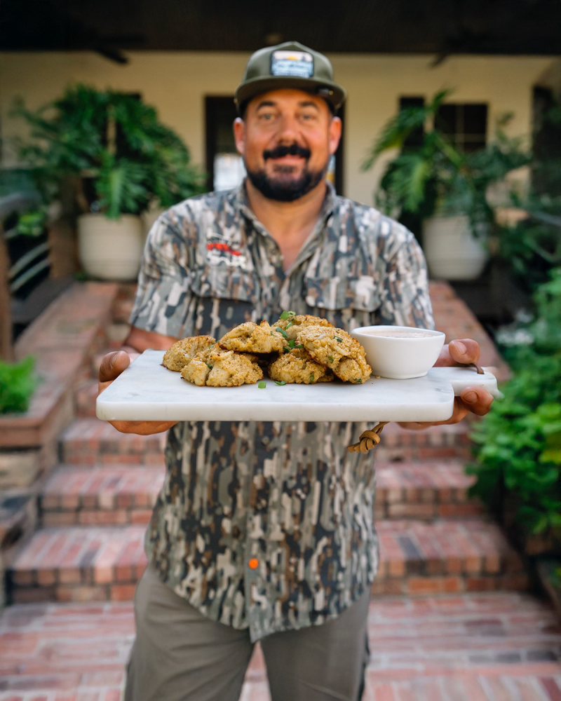 A man in camo stands on brick steps, holding a platter of Louisiana seafood and dipping sauce with plants behind him.
