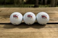 Three white golf balls with red and black logos lined up outdoors, ideal for Cajun cuisine or Louisiana crawfish enthusiasts.