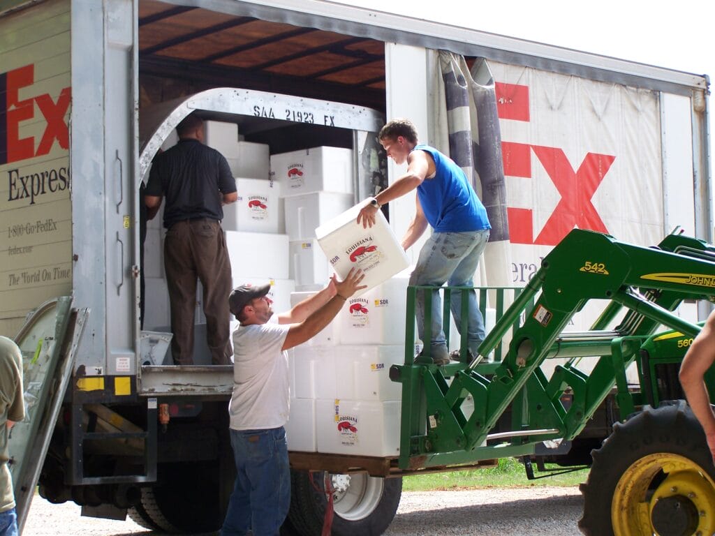 Three people use a tractor to unload fresh produce boxes from a truck, preparing Louisiana seafood for nationwide shipping.