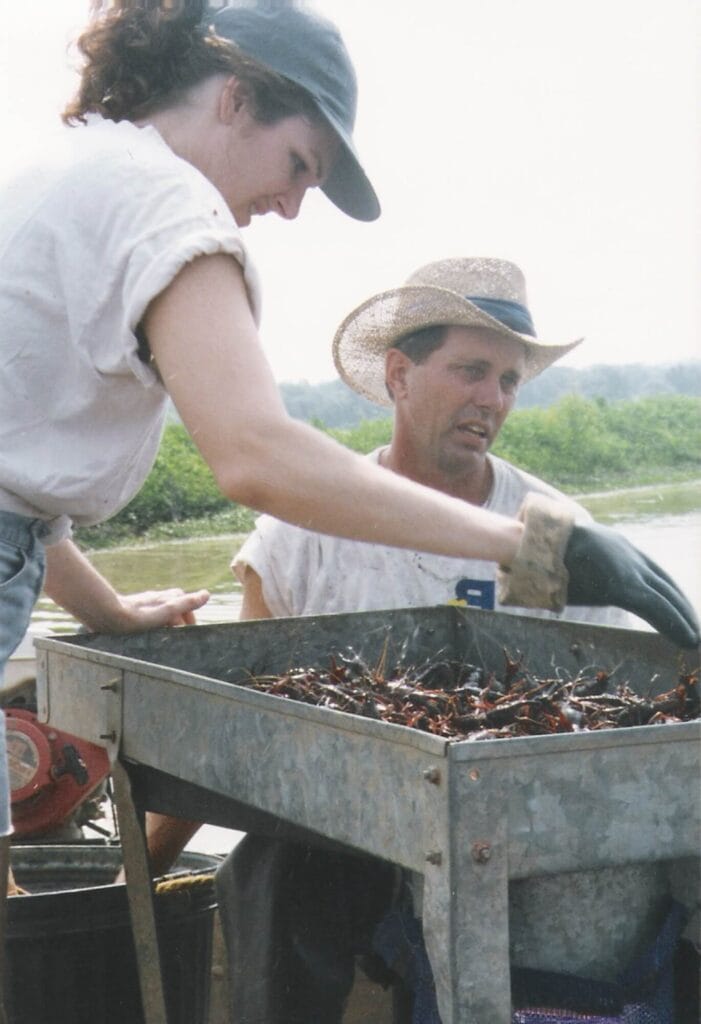 Two people sort Louisiana crawfish at a metal table outdoors, with lush greenery and water visible in the background.