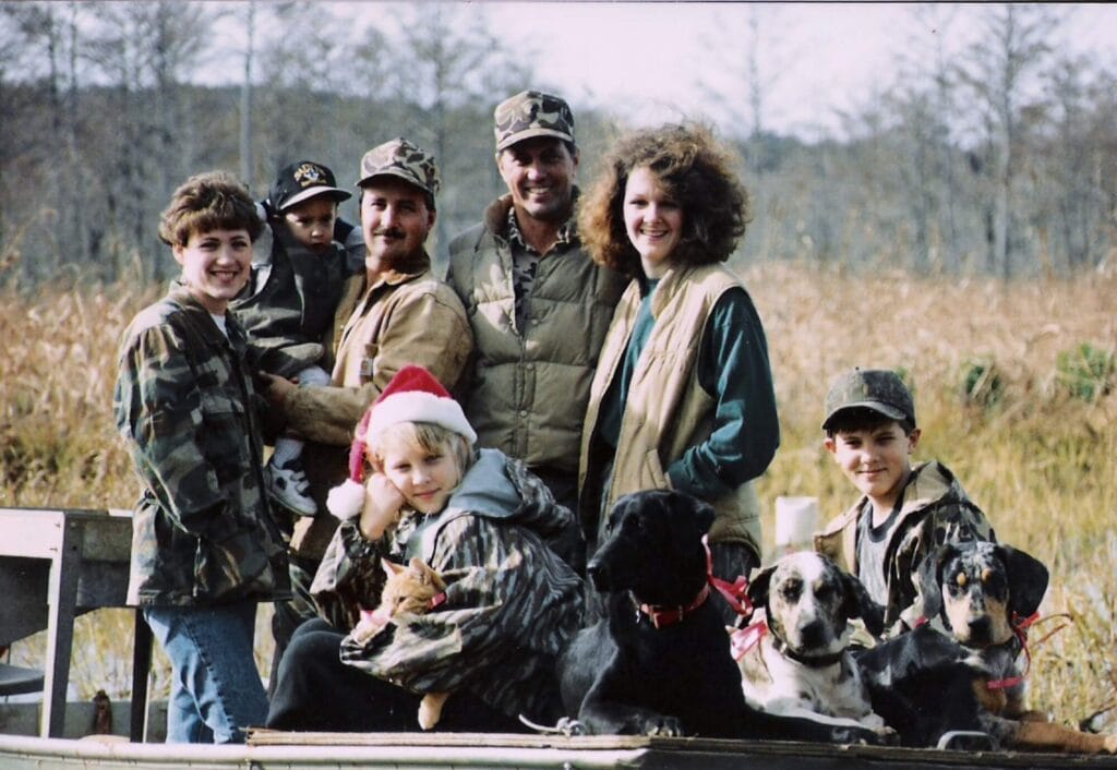 Seven people and three dogs pose on grass outdoors, some in camouflage, possibly dreaming of Louisiana crawfish or Cajun food.