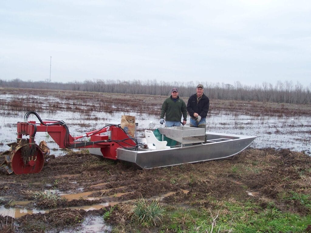 Two people harvest Louisiana crawfish in muddy fields beside a metal boat-like machine under a cloudy sky.