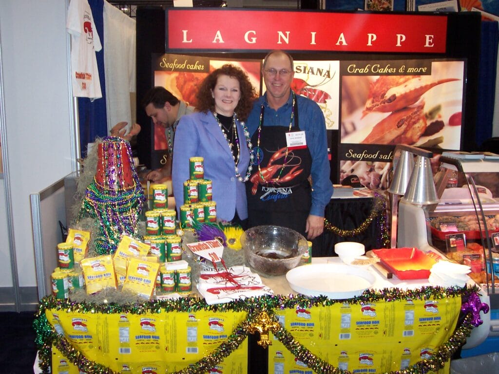 Two people stand behind a booth displaying Cajun foods and beads at a seafood trade show with Louisiana crawfish products.