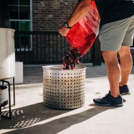 Outdoors, a person pours Louisiana crawfish into a metal basket beside a large pot for an authentic Cajun boil.