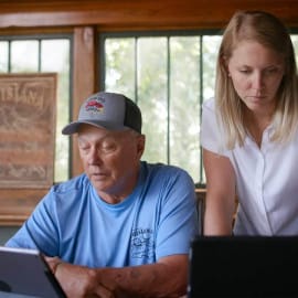 An older man and a younger woman browse devices on a table lit by sunlight, with Louisiana seafood and Cajun foods displayed.