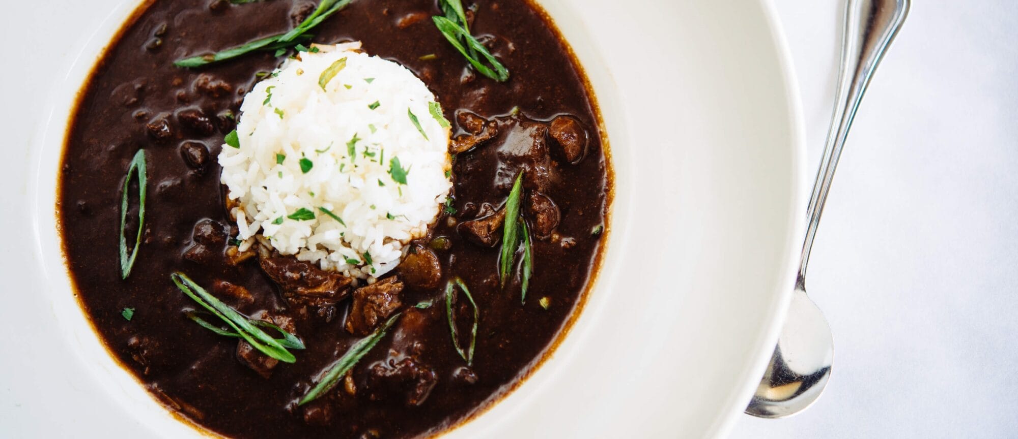 A bowl of dark Louisiana seafood stew with white rice and green onions sits next to a spoon, showcasing classic Cajun cuisine.