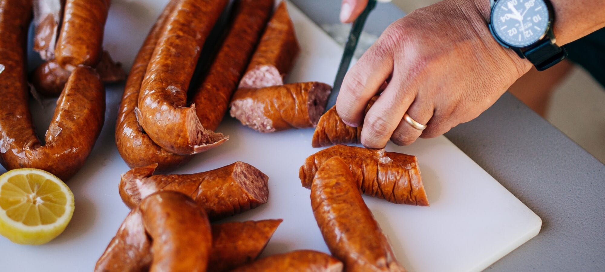 A person slices classic Cajun smoked sausages on a white cutting board, with a halved lemon, watch, and ring visible.