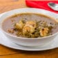 A bowl of LA Crawfish Co. Gumbo Sampler with a spoon and red napkin on a wooden table.