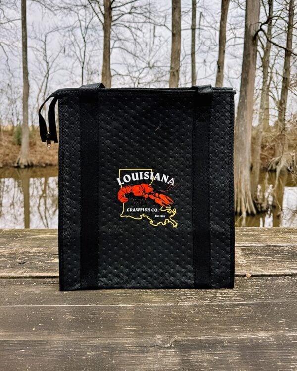 The LA Crawfish Co. Thermal Tote rests on wooden planks near a swamp, boldly displaying its logo and Louisiana crawfish heritage.