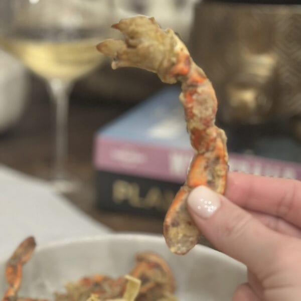 Pontchartrain Jumbo Lump Crab Swimmers 8 oz A hand holds an 8 oz Pontchartrain Jumbo Lump Crab Swimmers container, with blurred wine and books in the background.